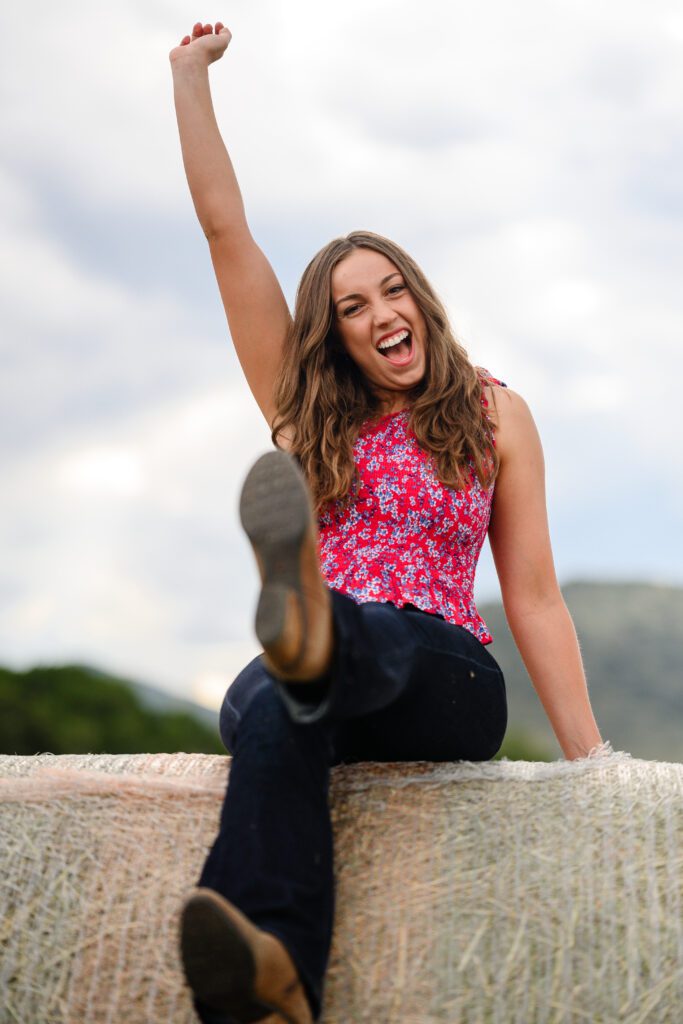 A girl celebrates with her arm up while sitting on a bale of hay and smiling at a Denver Senior Photographer!