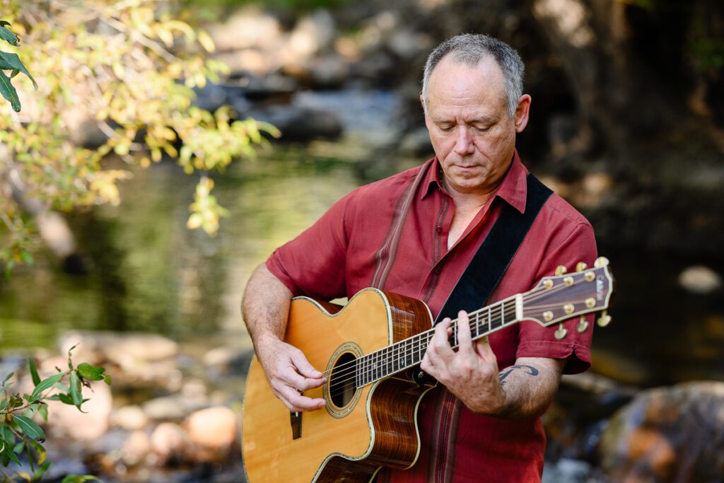 A closeup of a musician standing near a river bank playing guitar captured by a Denver branding photographer and brand strategist.