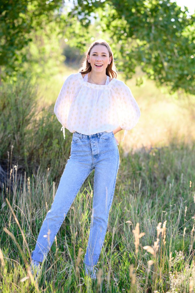 Standing in a green grass field with jeans and a white peasant shirt, a senior poses for her Denver Senior pictures.