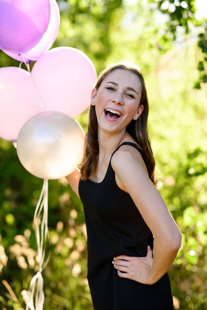 A denver senior photographer takes a picture of a cute girl in a black dress laughing at the camera while holding a bunch of purple balloons as a fun prop for her senior photos.