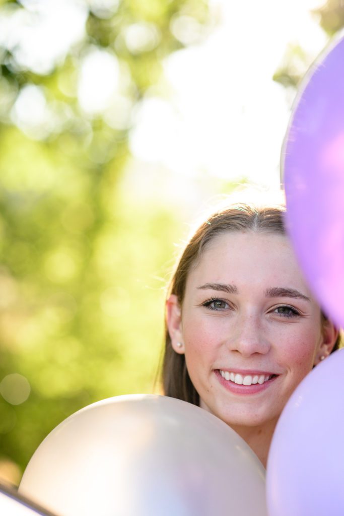A closeup of a girl smiling at a Denver senior photographer for her senior photos with purple balloons all around her face.
