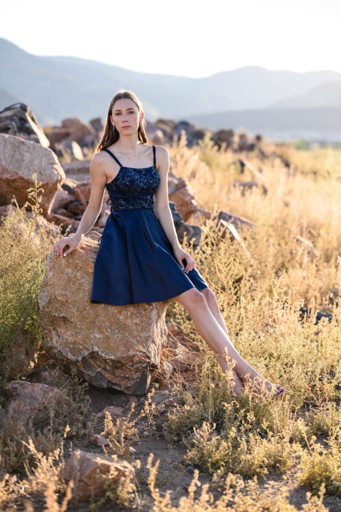 A girl in a blue dress looks like a model as she lounges on a rock in front of a beautiful Colorado mountain backdrop for a Denver Senior Photographer at Bear Creek Lake Park.