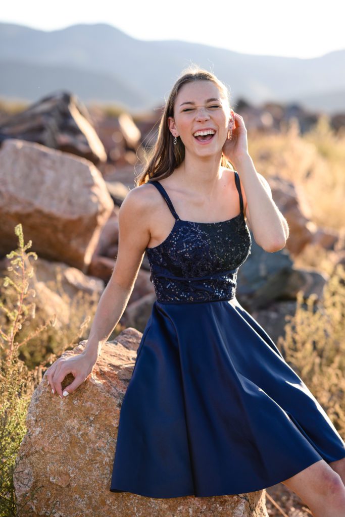 A brightly lit senior picture for a girl in a blue fancy dress sitting on a rock in front of an iconic Colorado mountain backdrop smiling at a Denver Senior Photographer.