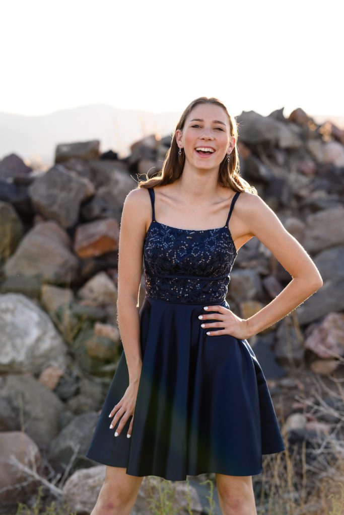 A Denver senior photographer takes a picture of a girl in a fancy blue dress standing in front of a large pile of rocks with a Colorado mountain background.