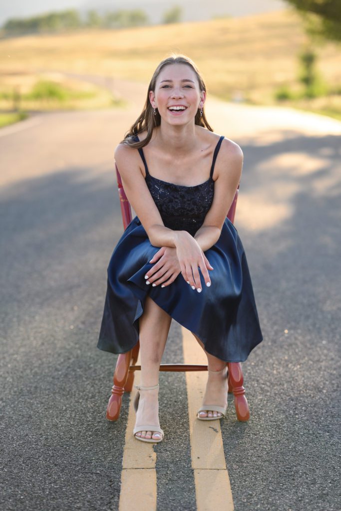 A denver senior photographer captures a classic Colorado background with a girl in a blue fancy dress sitting on a red chair in the middle of a road for her senior pictures.