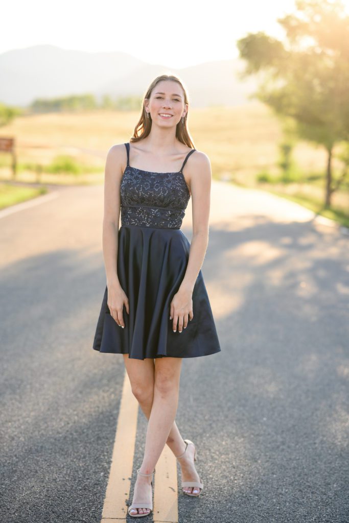 A girl in a blue sparkly dress standing in the middle of a road with mountains in the background smiling at a Denver Senior Photographer.