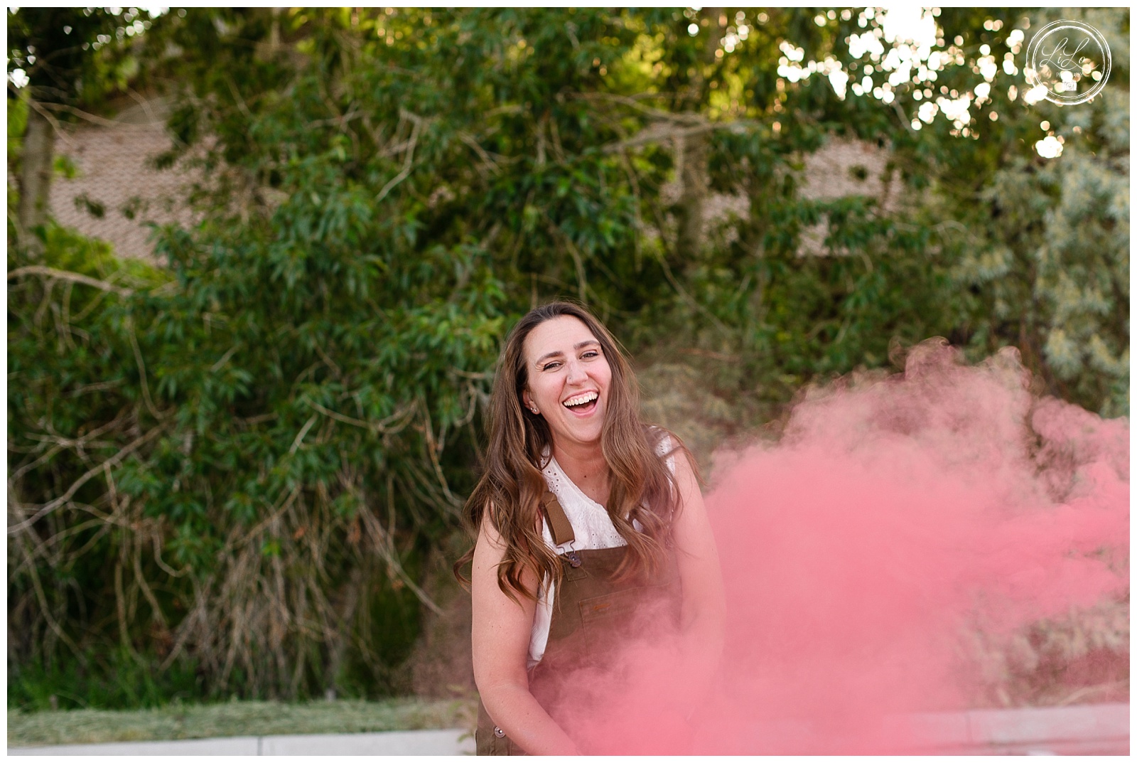 Denver senior photographer captures outdoor senior photos with female model laughing as a pink smoke bomb goes off in front of her