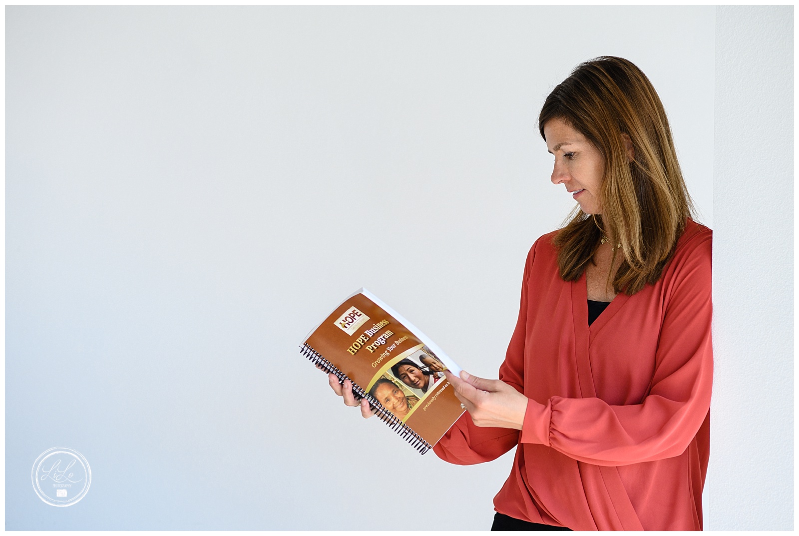 Denver Brand Photographer captures a woman in a salmon colored shirt standing in front of a white wall looking at a brown workbook.