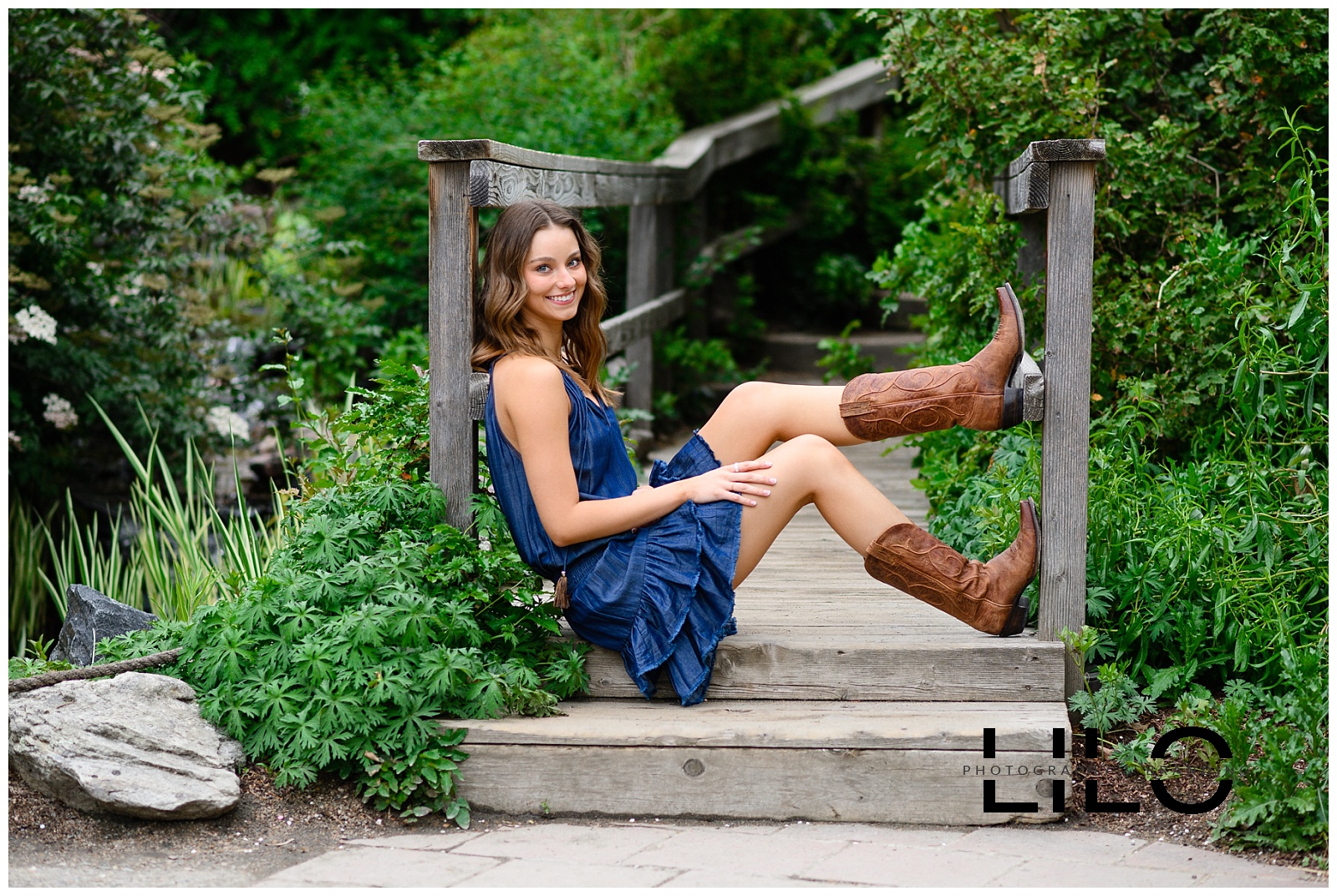 Denver Senior Pictures at Denver Botanic Gardens with young woman sitting on a bridge while wearing a dark blue dress and boots photographed by Denver senior photographer