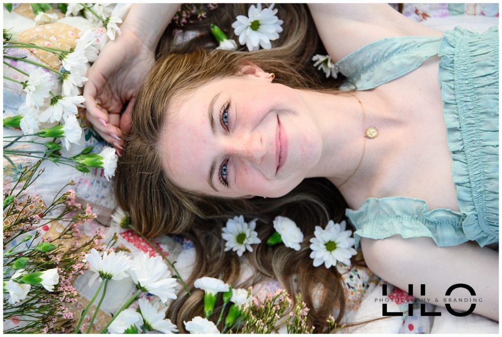 A closeup image of a girl with green eyes laying on a blanket in a green sleeveless top and flowers all round smiling at a Denver Senior Photographer.