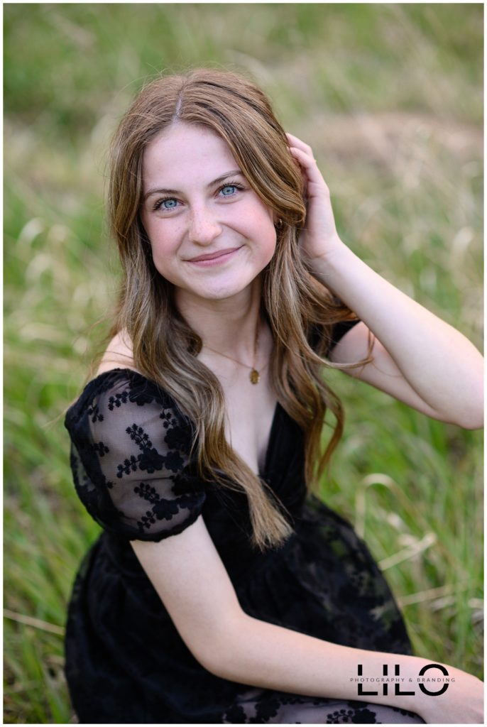 Denver senior photos of a girl with long blonde hair and striking green eyes sitting in the grass with a black dress on.