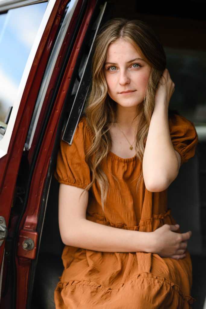 Denver Senior Photographer takes a picture of a serious green eyed senior wearing an orange dress sitting in an old VW van.