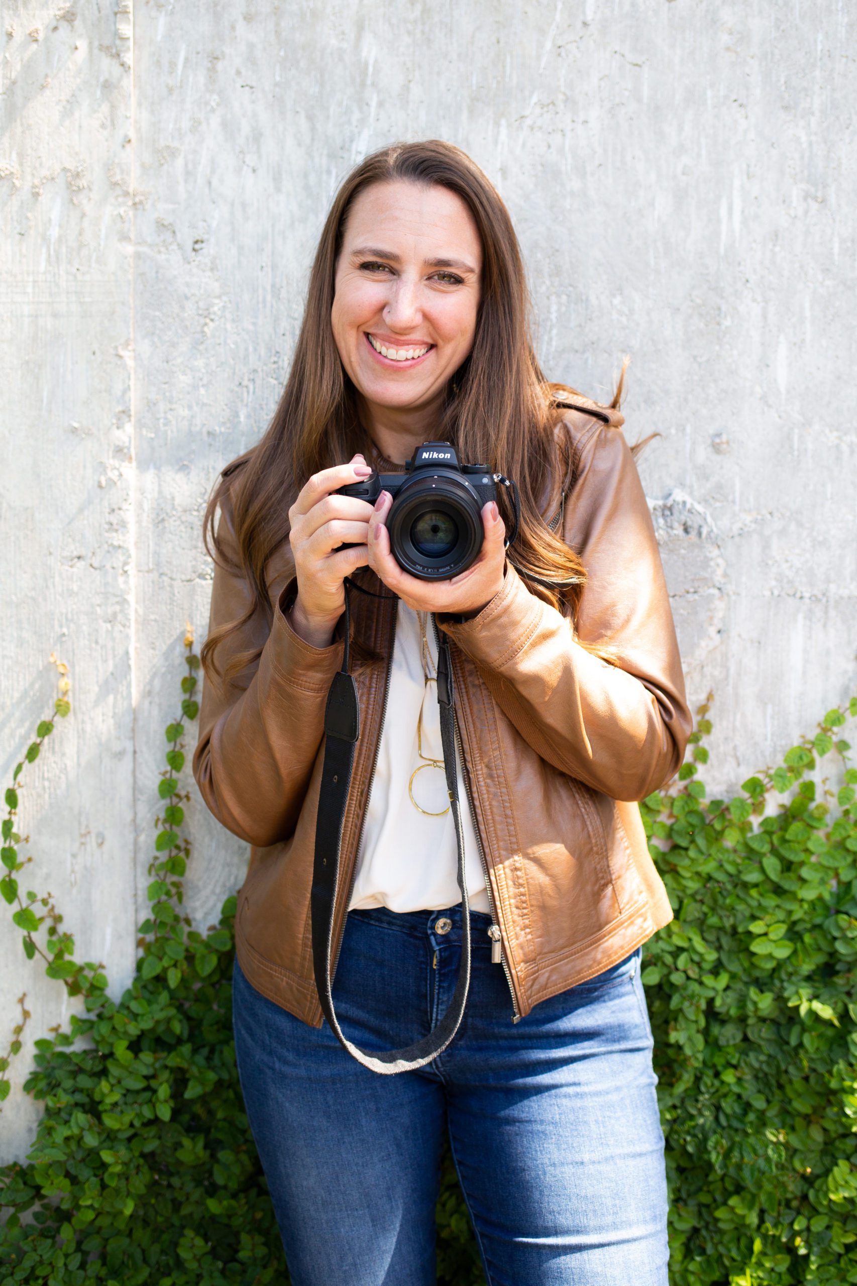 A woman in a brown leather jacket is holding a nikon DSLR camera and smiling at Denver Commercial Photographers.