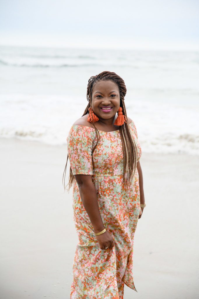Black woman with long corn rows in a bright orange dress and earrings posing on a beach with the ocean in the background for Denver Commercial Photographers.
