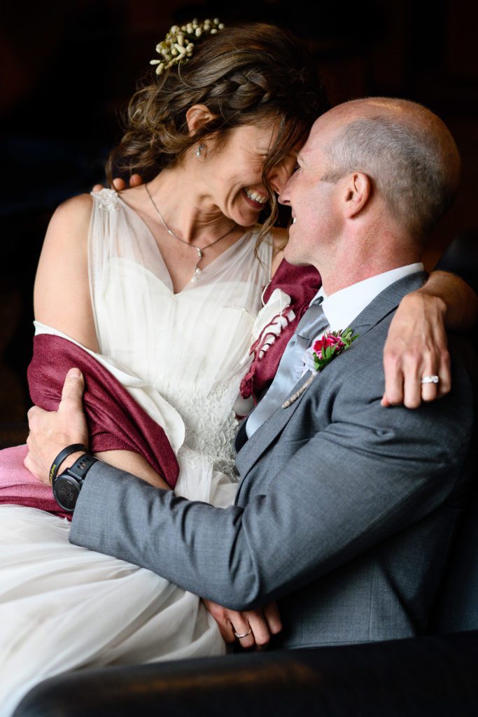 Bride smiling while sitting on the lap of her groom as he has his arms wrapped around her as captured by a Denver Photographer.