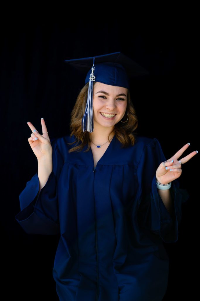 Girl giving peace signs in a blue graduation cap and gown smiling at a Denver Senior Photographer.
