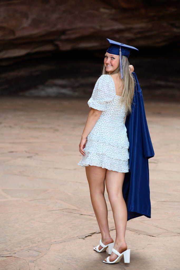 Denver Senior Photographer takes picture of a young lady with long blonde hair walking away from the camera with her blue graduation gown draped over her shoulder.