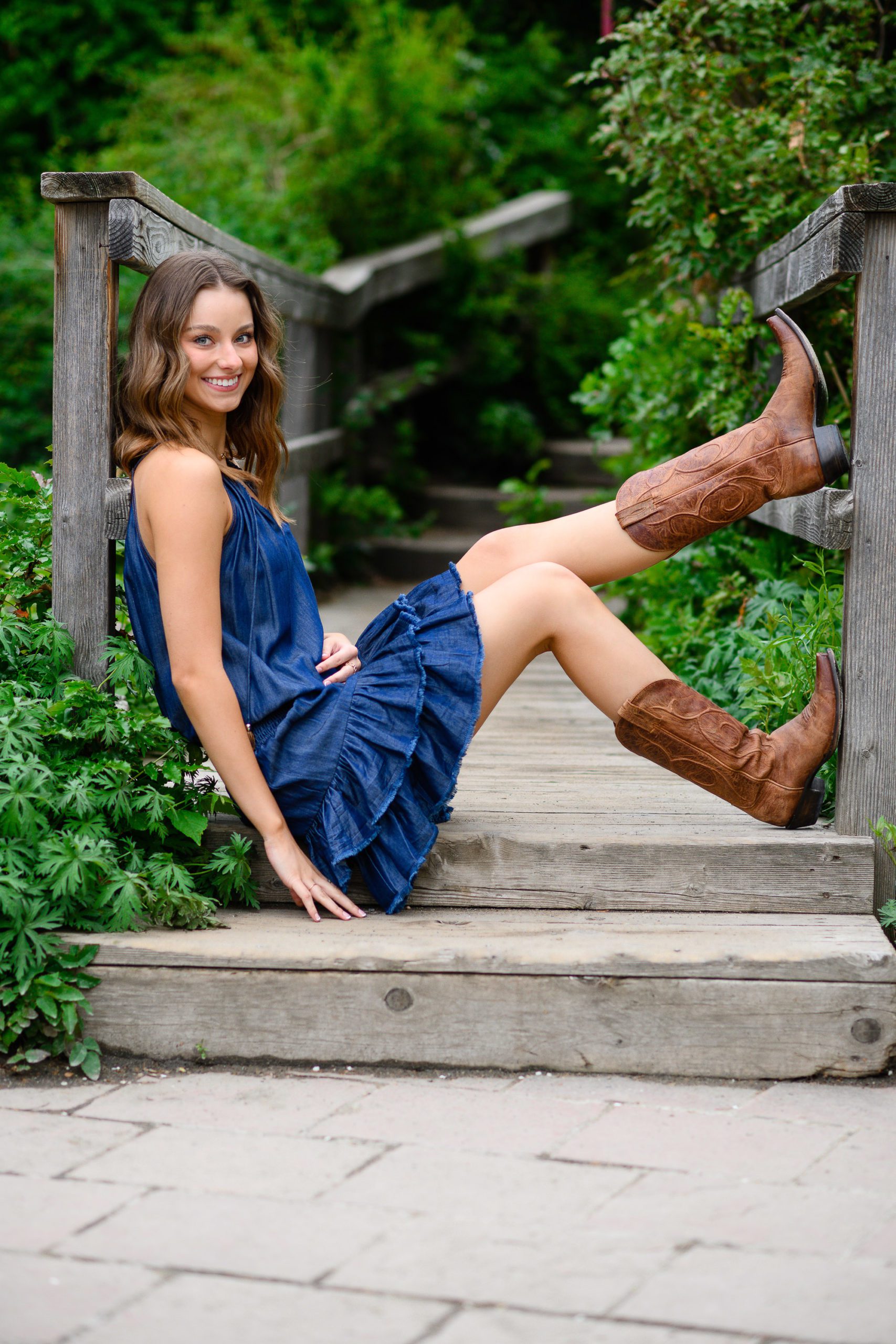 Senior girl in a blue denim dress and cowboy boots sits at the edge of a bridge with her feet kicked up for a Denver Senior Photographer.