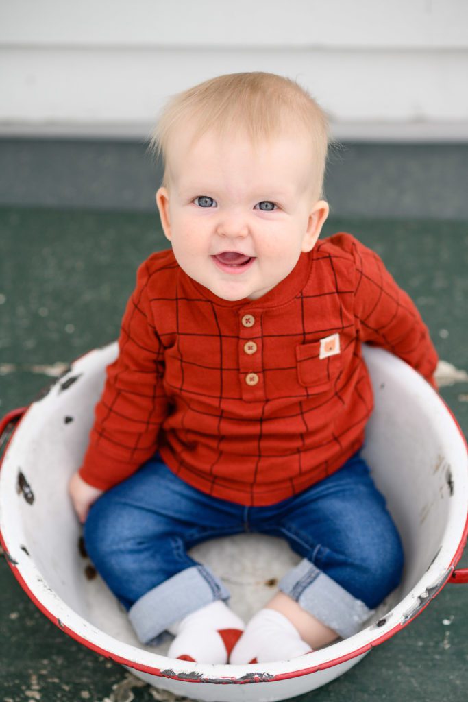 8 month old baby boy in jeans and an orange long sleeved henley shirt sitting in a vintage white pot with red handles looking at a Denver Photographer and grinning.