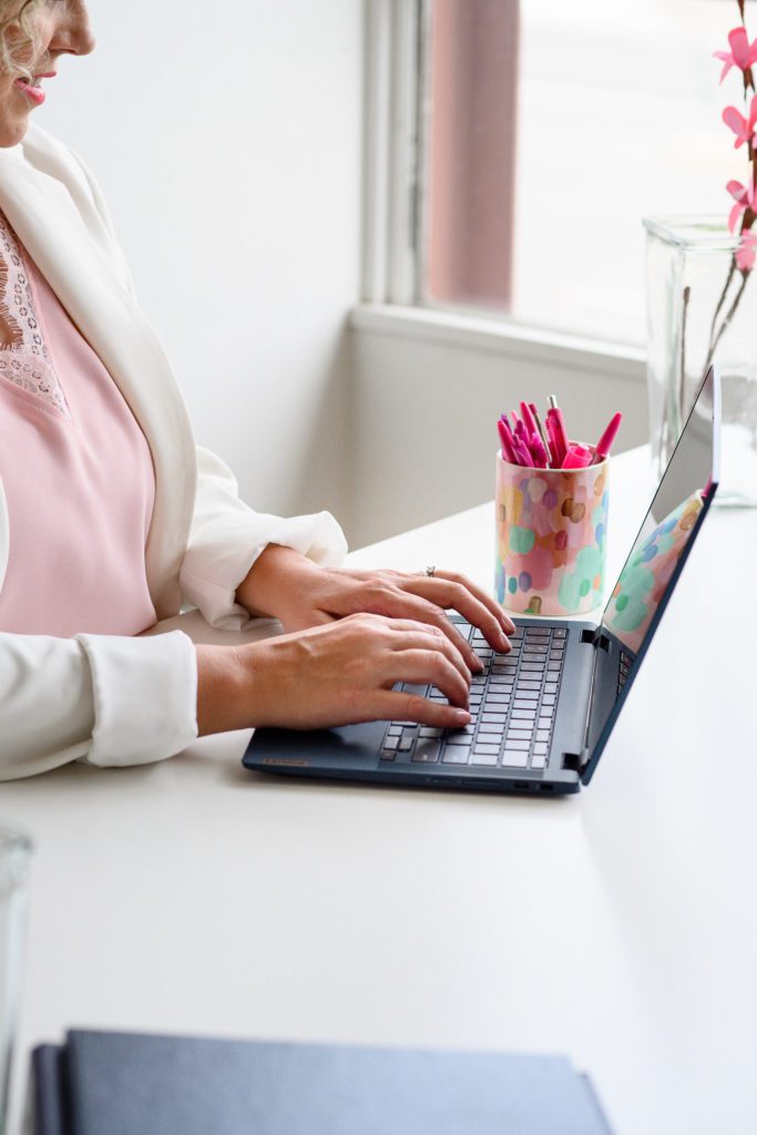 Denver Commercial Photographers capture a woman sitting in a white office with a white blazer and pink shirt typing on a black laptop with brightly colored pens in the background.