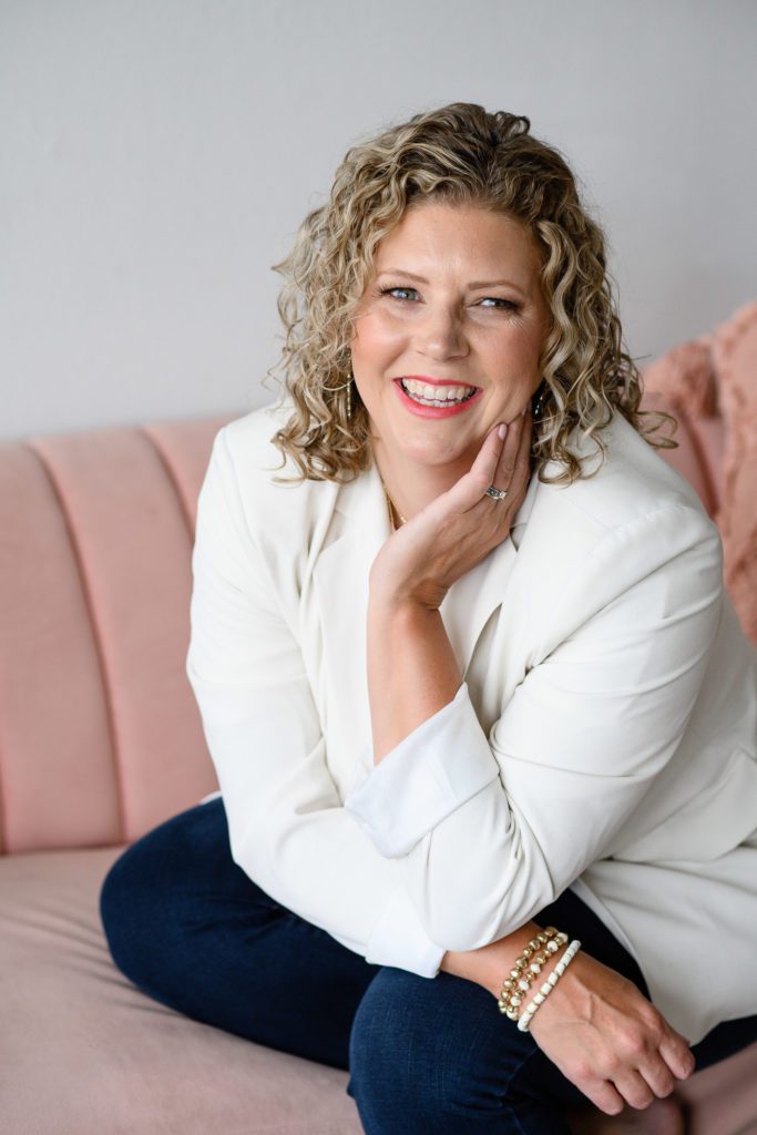 A woman in a white blazer sits smiling at a Denver Commercial Photographers on a pink couch with her left arm up near her neck.