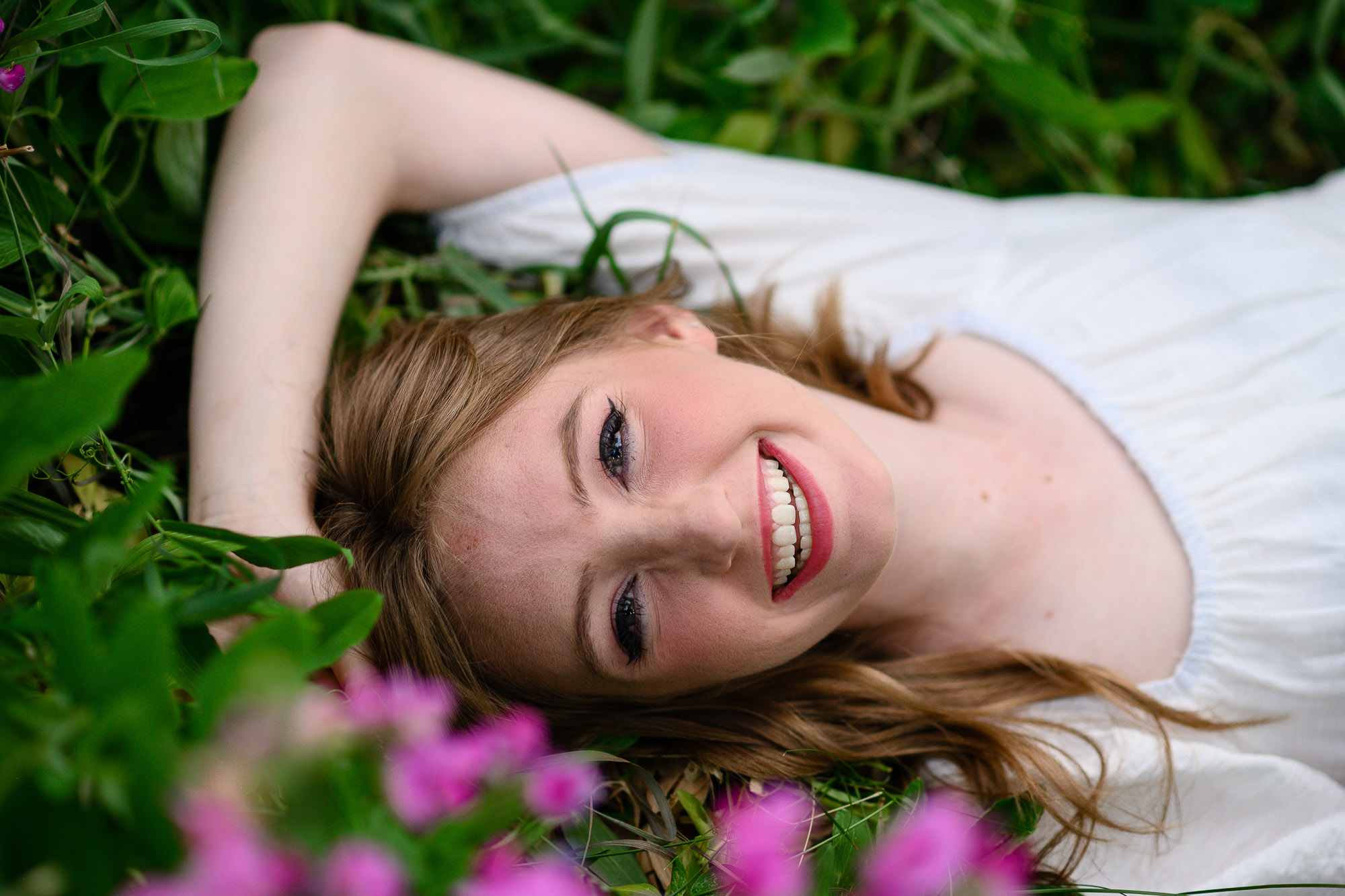 Senior girl laying in a field of pink flowers smiling at a Denver Senior Photographer with her hand over her head.