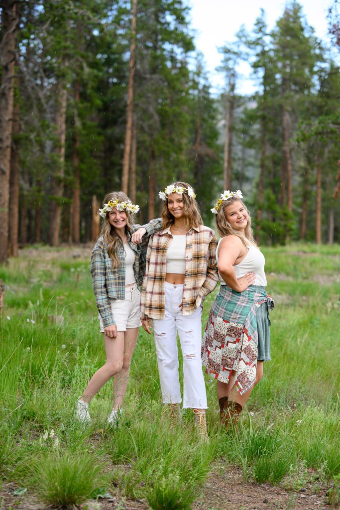 Three senior girls stand in a mountain meadow with flower crowns on and flannel shirts leaning on each other and smiling at a Denver Senior Photographer.