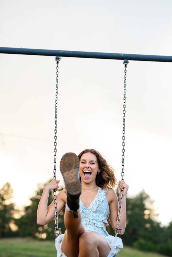 Denver senior photographer takes a picture of a senior girl on a swing set with her boot outstretched to the camera and a big smile on her face.