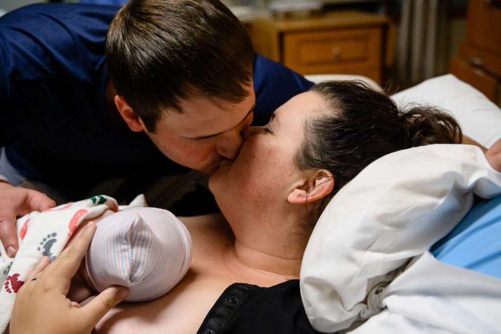 Man and woman kiss each other in a hospital with their newborn baby on the woman's chest captured by a Denver Photographer.