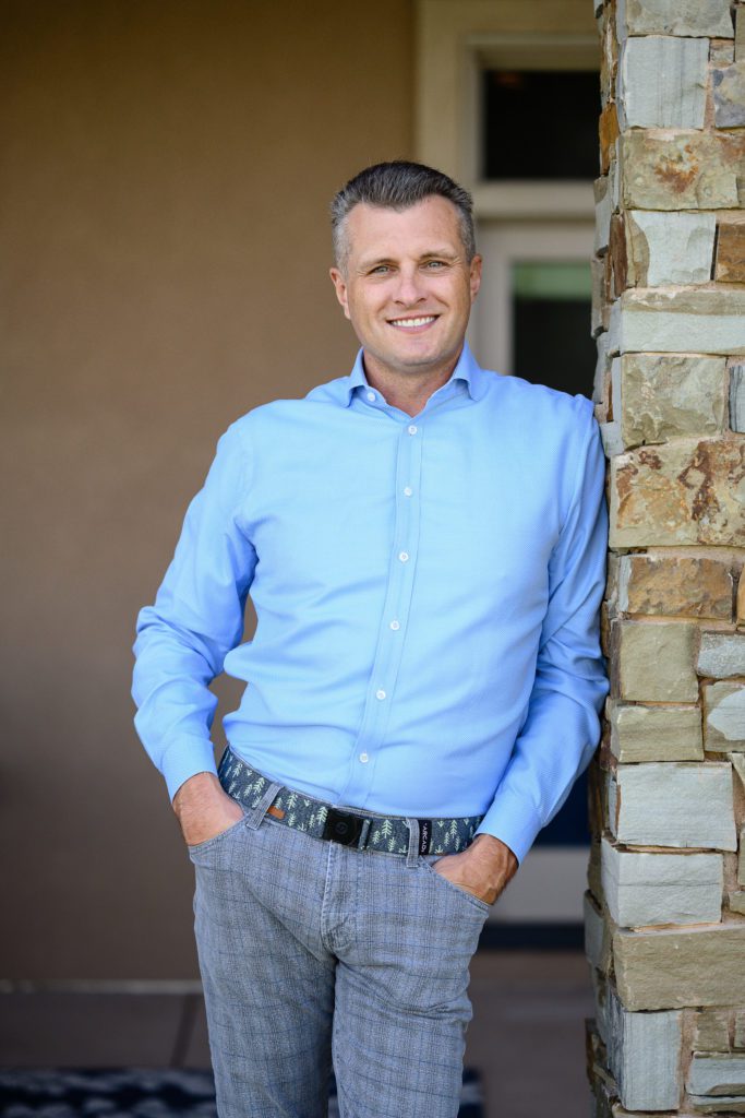 Man in bright blue dress shirt leans on a stone column with his hands in his pockets and smiles at Denver Commercial Photographers.