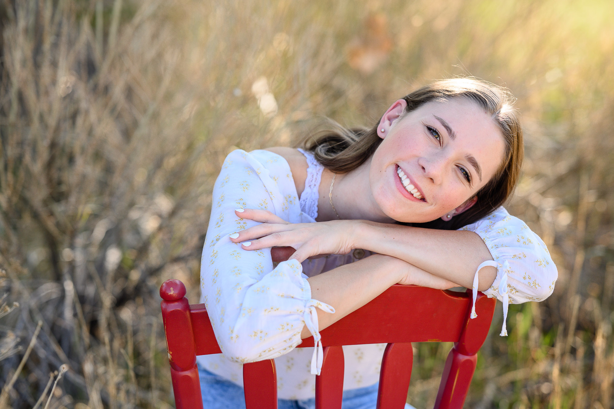 Senior girl sits backwards in a red chair and leans her head and arms on the back of the chair and smiles at a Denver Senior Photographer.