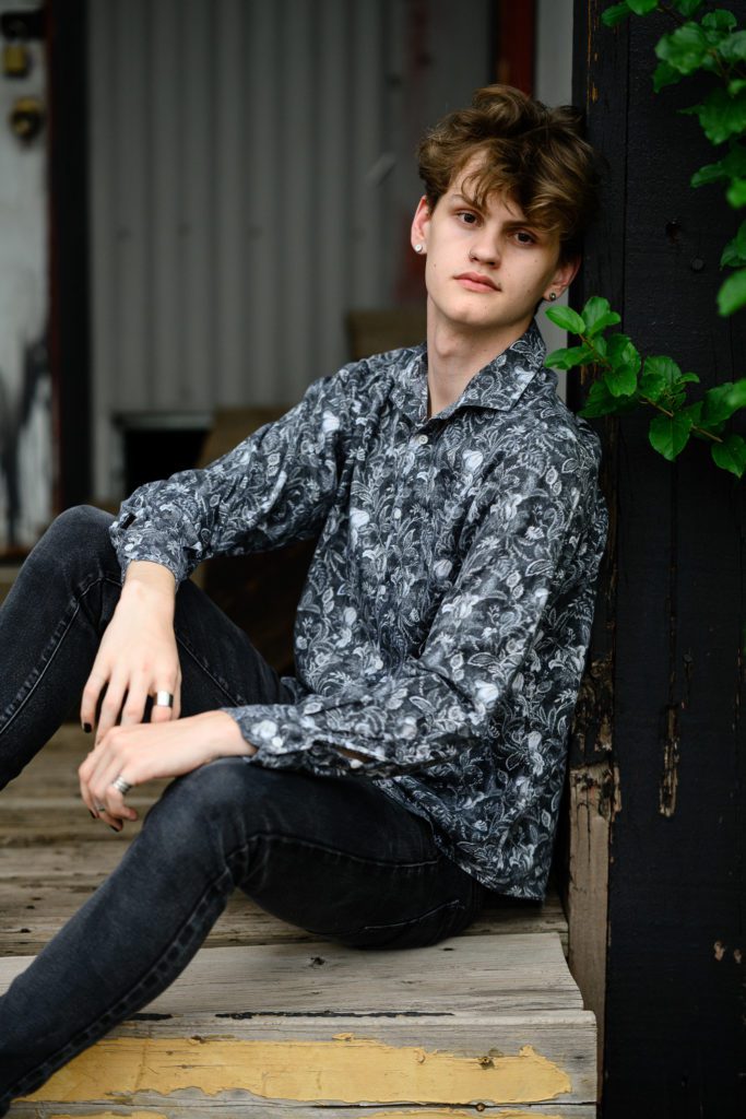 A goth looking boy in a black paisley shirt and jeans with black fingernails leans against an old building while sitting on a set of stairs looking straight faced at a Denver Senior Photographer.