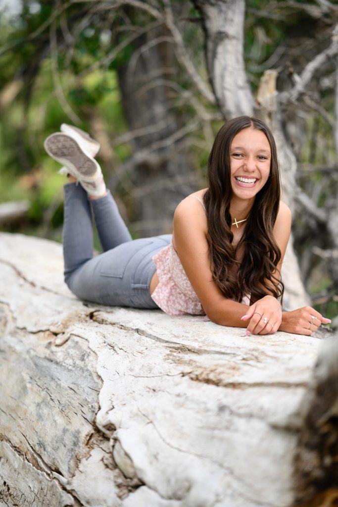 Young senior lays on her belly on an old bare tree smiling at a Denver Senior Photographer.