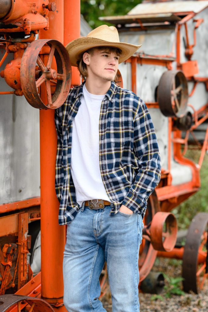 A senior boy in a flannel shirt, jeans and a cowboy hat leans against an old metal and orange piece of machinery looking away from a Denver Senior Photographer.