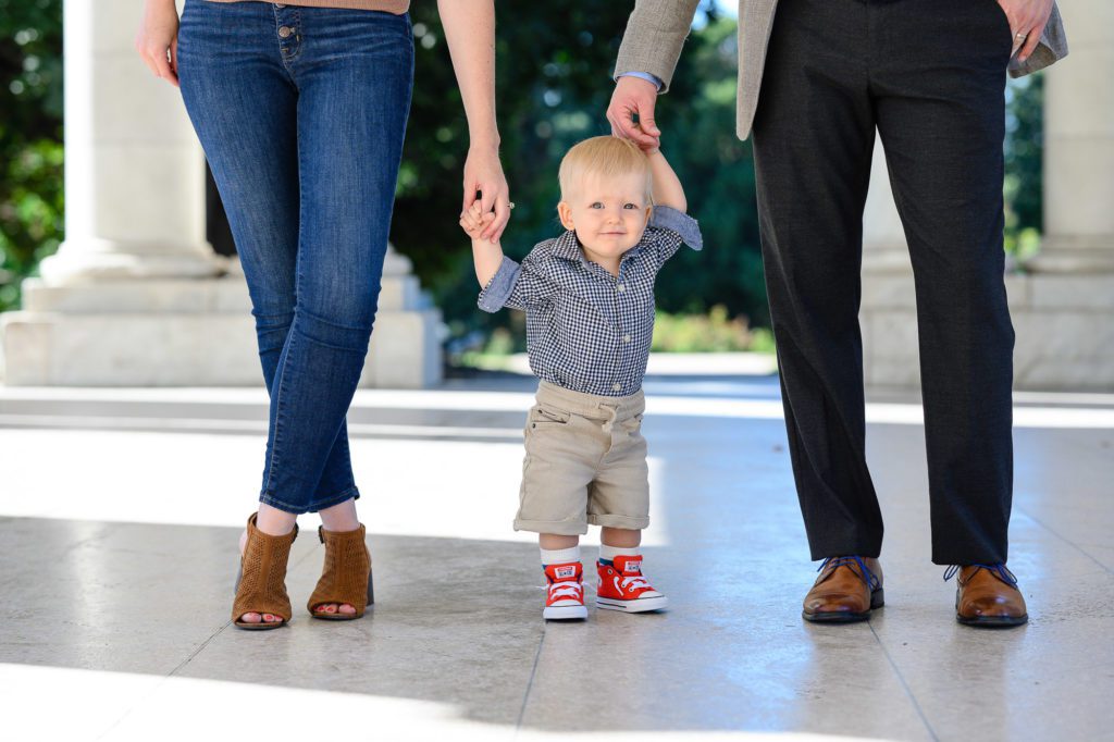 A one year old boy in red converse, khaki shorts and a denim shirt holds hands with his mom and dad while smiling at a Denver Photographer.