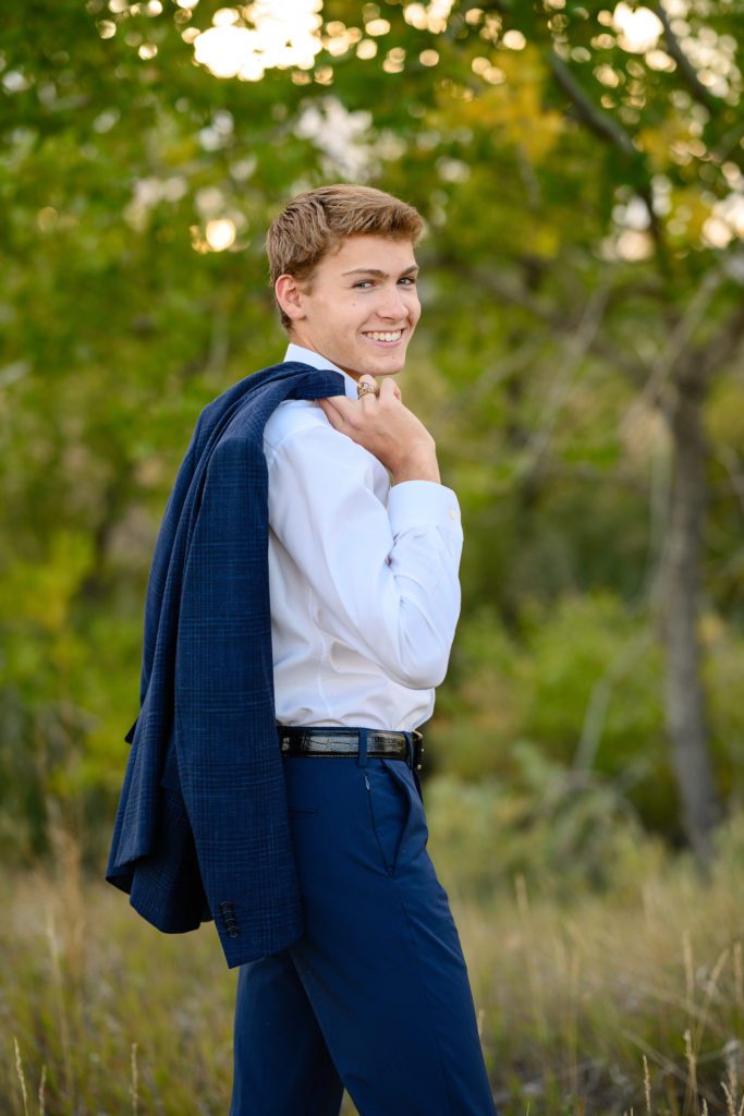 A senior boy in a blue suit looks back over his shoulder with his suit jacket flung over his shoulder grinning at a Denver Senior Photographer.