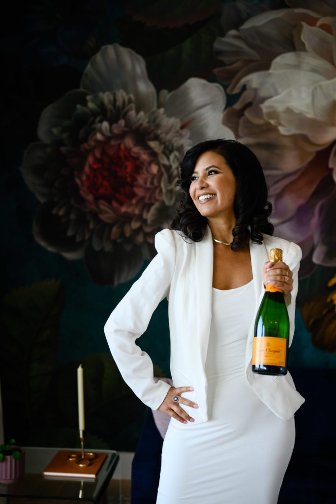 A Latina woman in a formal white business suit holds a bottle of champagne toward Denver commercial photographers in a room with giant flowered wallpaper.