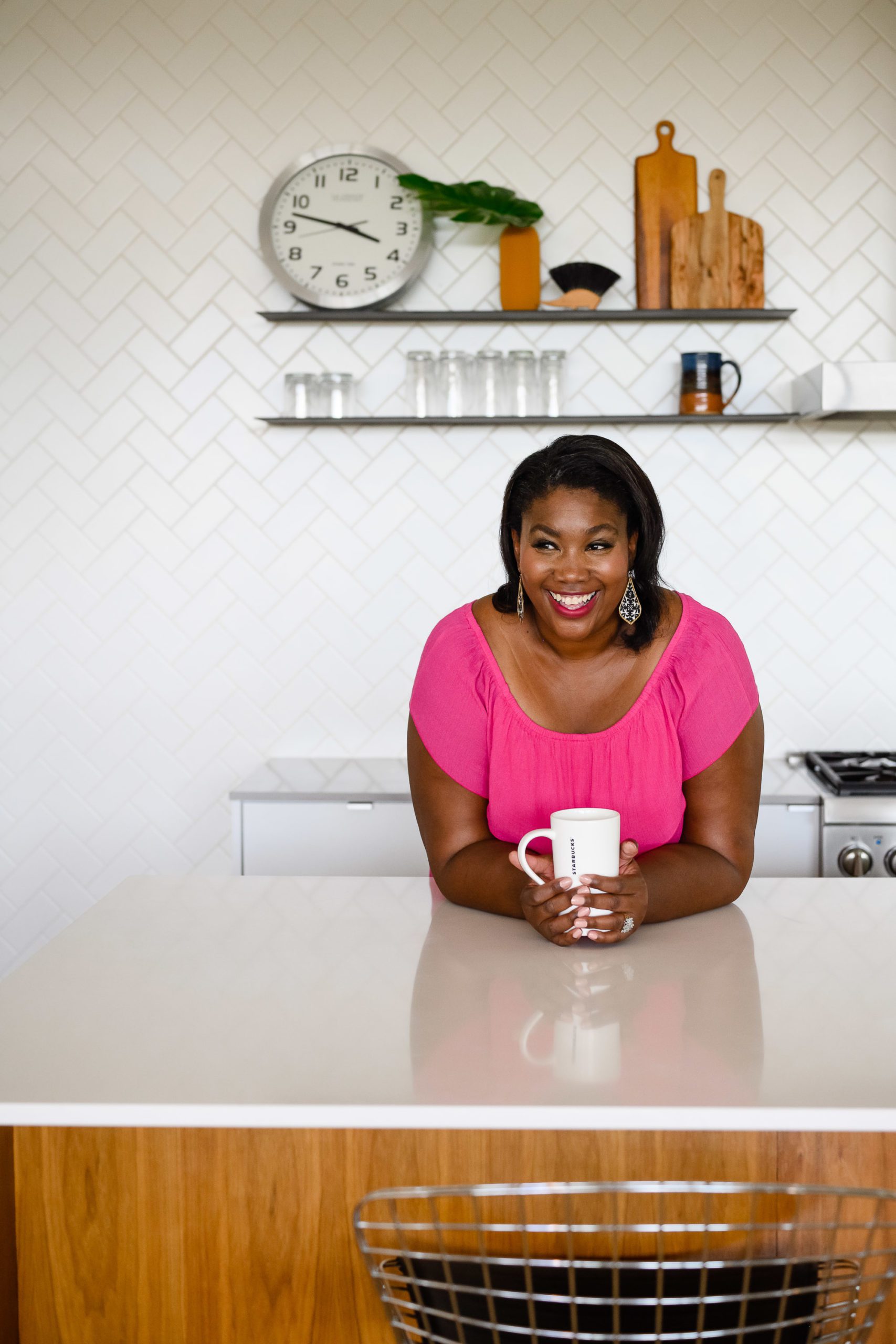 A black woman in a bright pink dress hold a white starbucks coffee mug and smiles away from Denver Commercial Photographers while in a white, modern kitchen