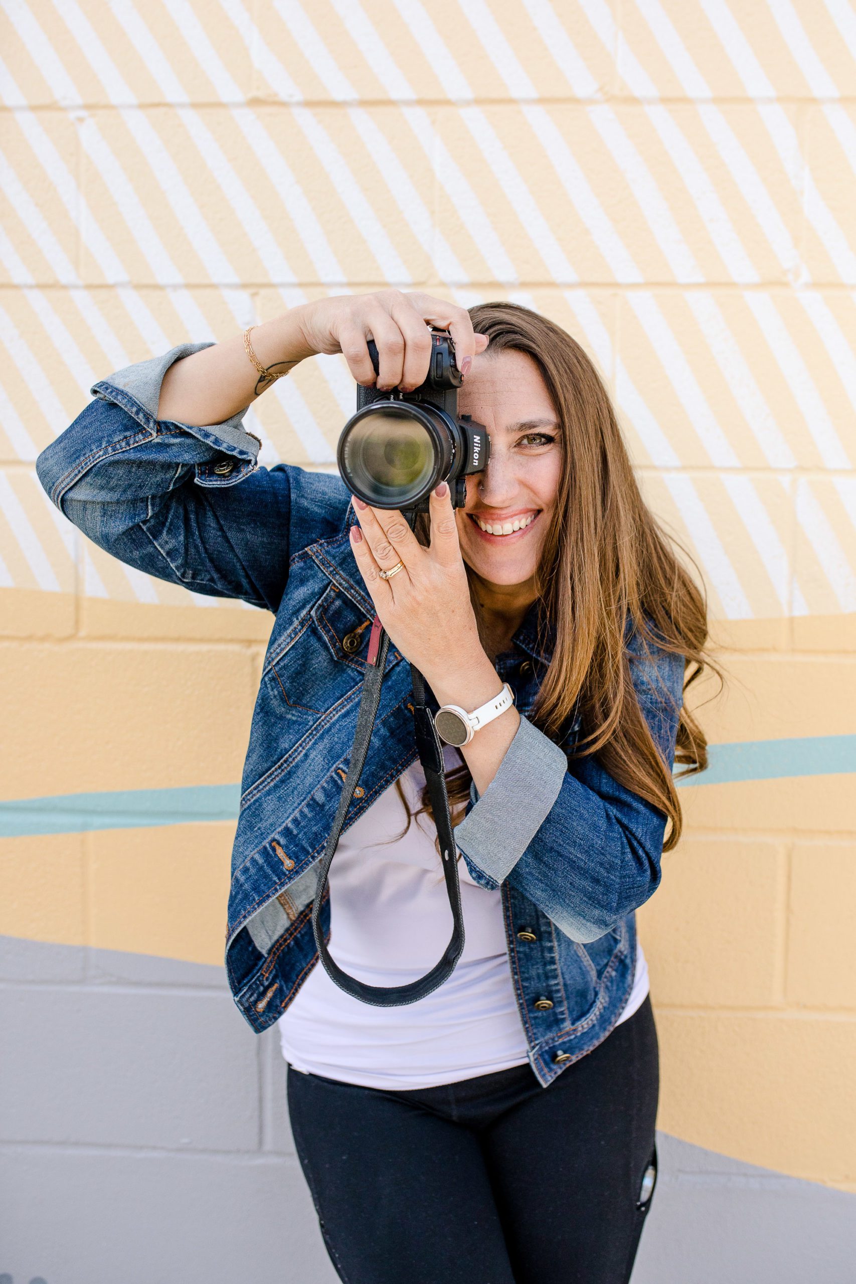 Woman standing in front of a yellow striped wall wearing a white shirt and jean jacket holds up a camera to her face and smiles at Denver commercial photographers.