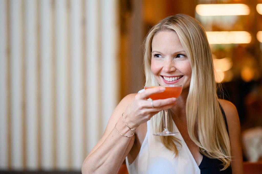 A blonde woman in a nice white dress sits at a bar and holds a pink colored martini and smiles away from Denver commercial photographers