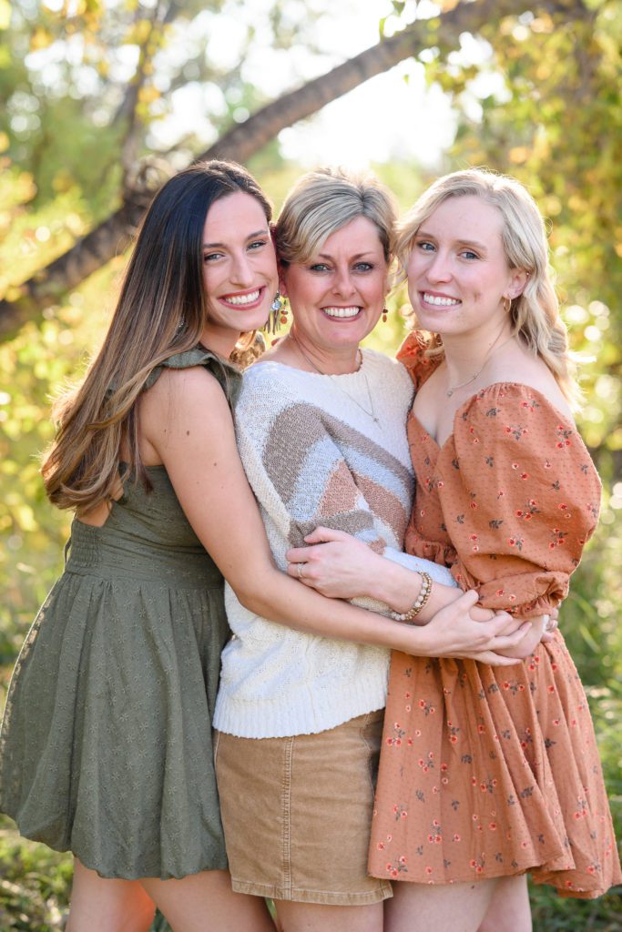 Two grown sisters in fall colored dresses hug with their mom in the middle captured by a Denver Photographer.