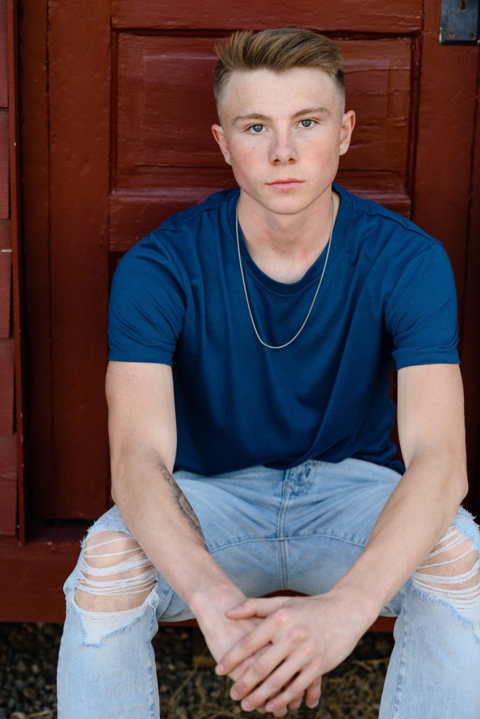 Senior boy sits with a serious face toward a Denver Photographer in a blue t-shirt in front of a red door.
