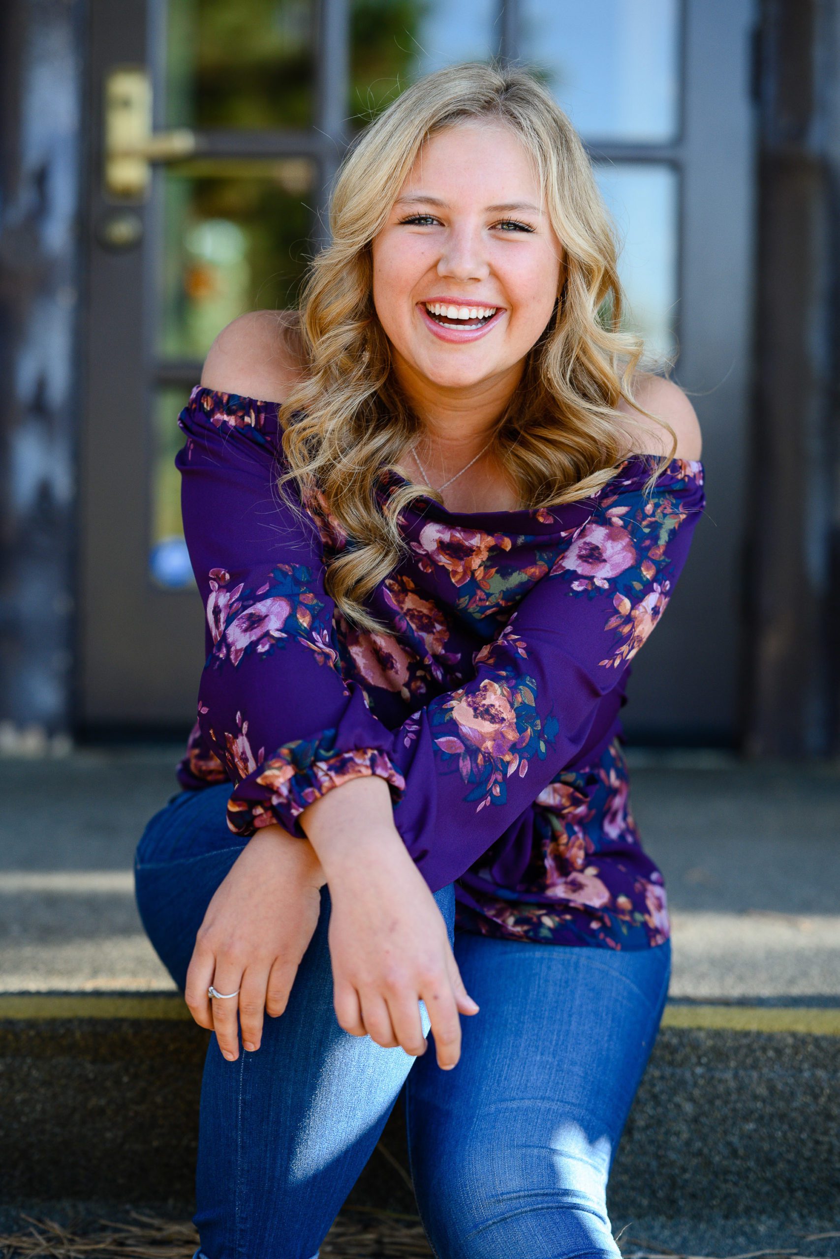 Denver Photographer takes a picture of a blonde senior with big curls in a purple off the shoulder shirt sitting on stairs.