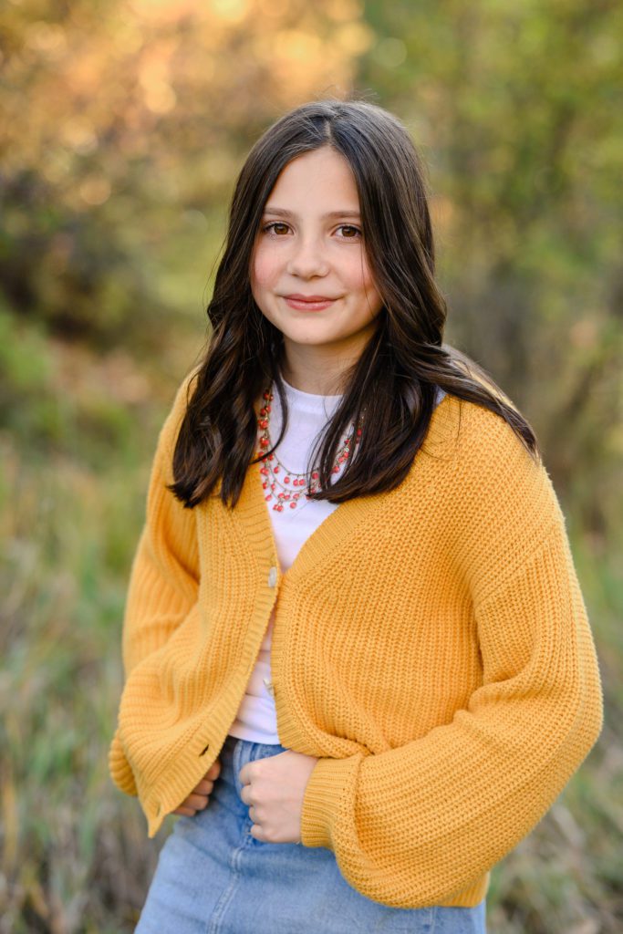 Tween girl in a yellow sweater with amber eyes lightly smiles at a Denver photographer with fall leaves blurred in the background.