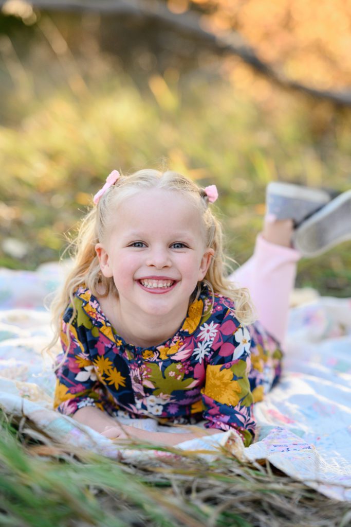 Little blonde girl in a flowered top lays on her tummy on an old quilt smiling at the camera.