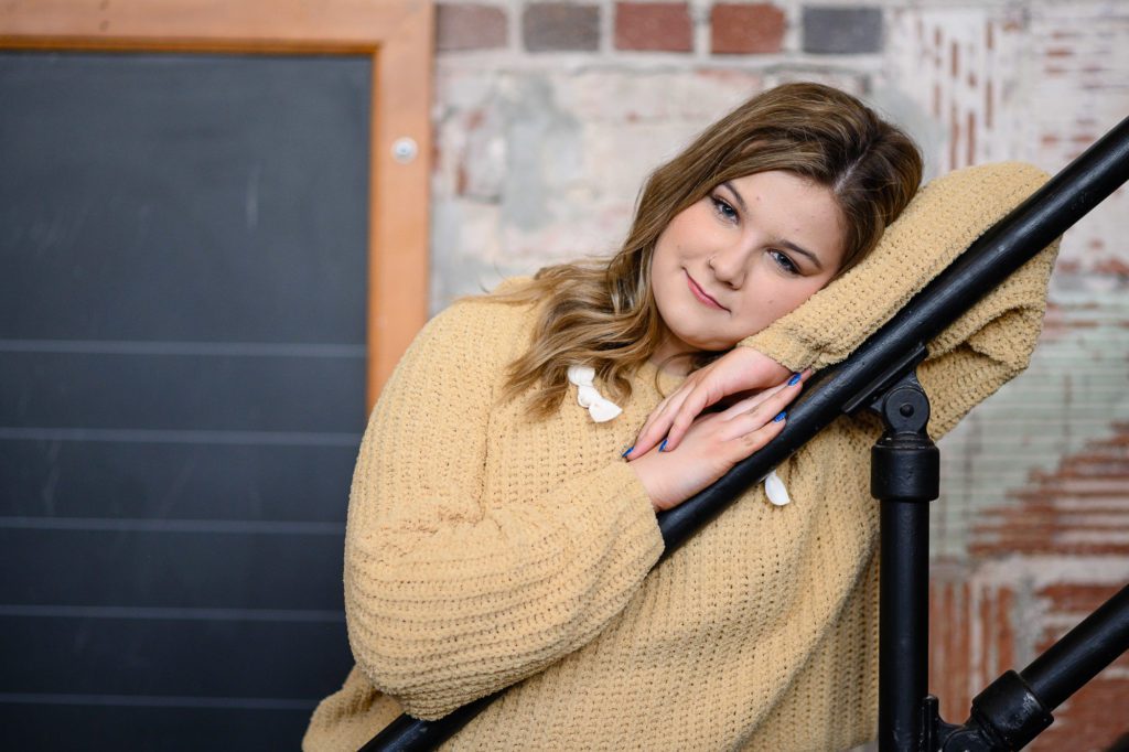 A young girl in a yellow sweater with her head resting on a staircase railing with a soft smile towards a Denver Senior Photographer.