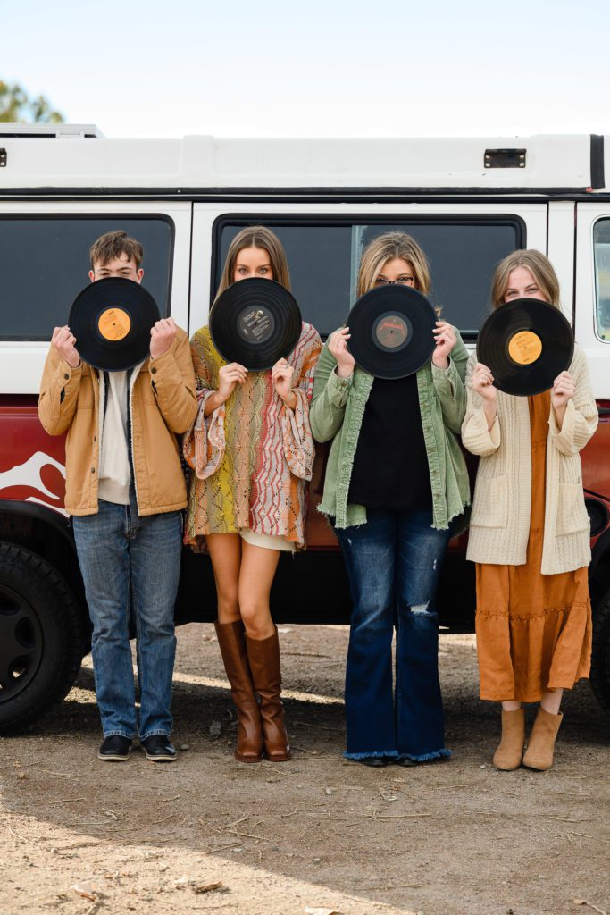 Four teenage seniors wearing 70's clothes hold vintage records in front of their faces in front of a vintage VW westfalia van captured by a Denver Senior Photographer.