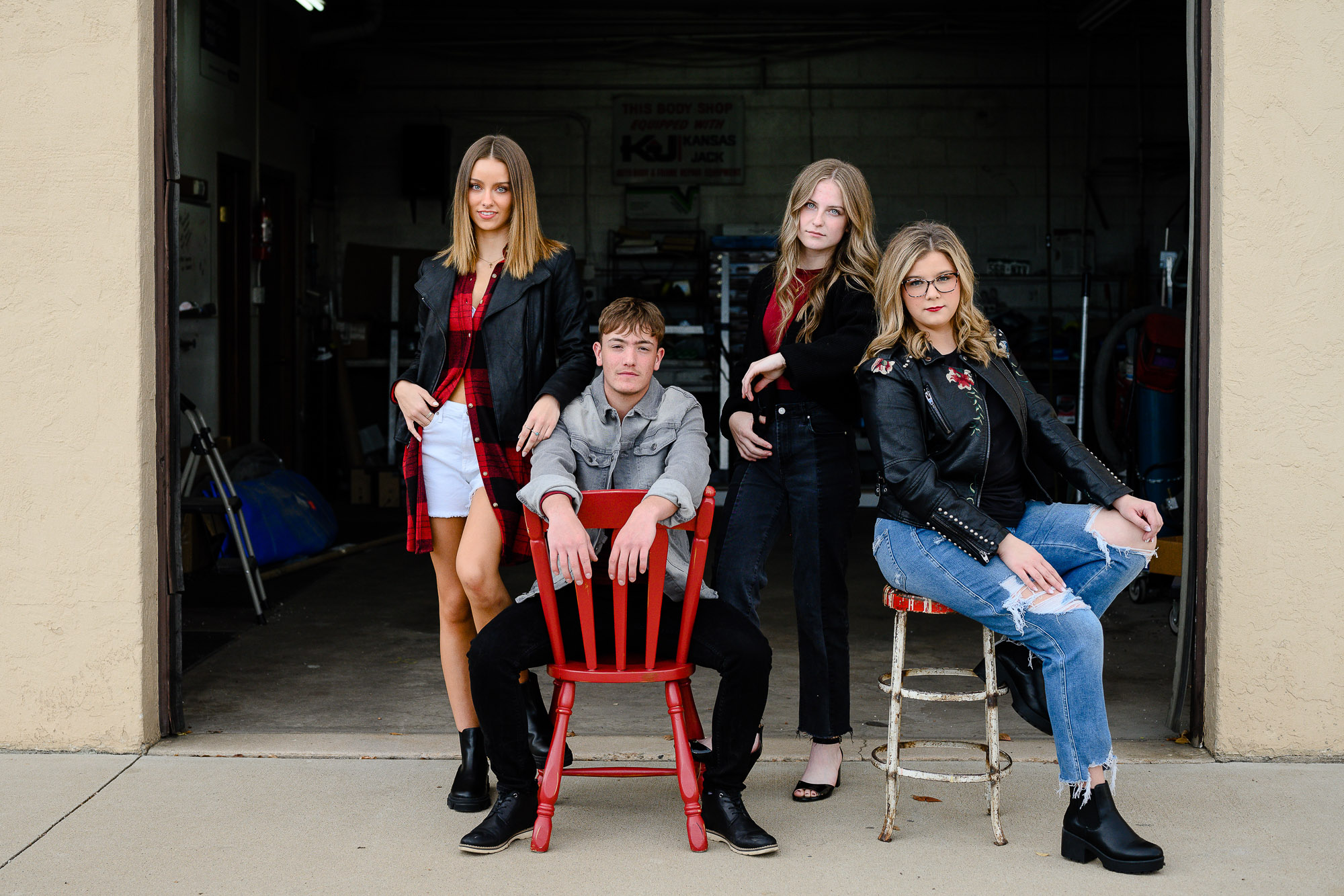 Four teenage seniors in black and red clothes sit and stand in front of an open auto body garage with serious looks on their faces while looking at a Denver Senior Photographer.