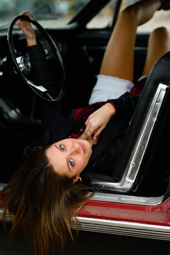 Senior Girl laying in a vintage red car with her back on the front seat holding the steering wheel and gazing at a Denver Senior Photographer.