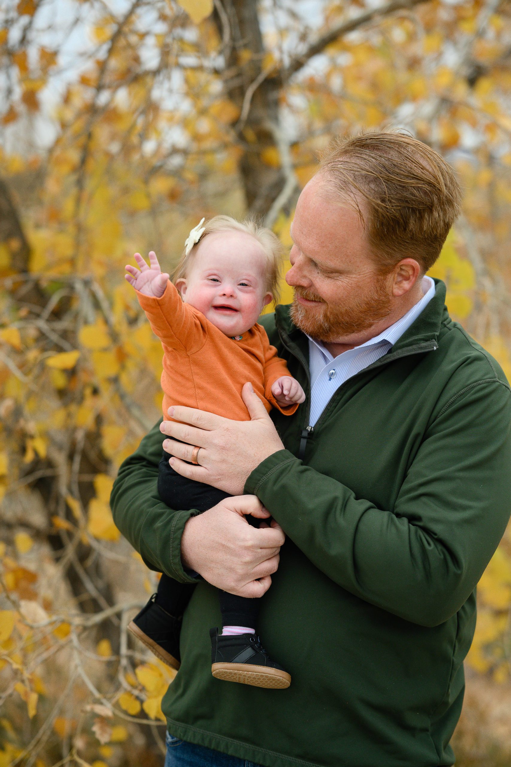 A Denver Photographer catches a little girl with down syndrome in a bright orange shirt smiling and waving at the camera while her dad in a green sweater is holding her and smiling at her.